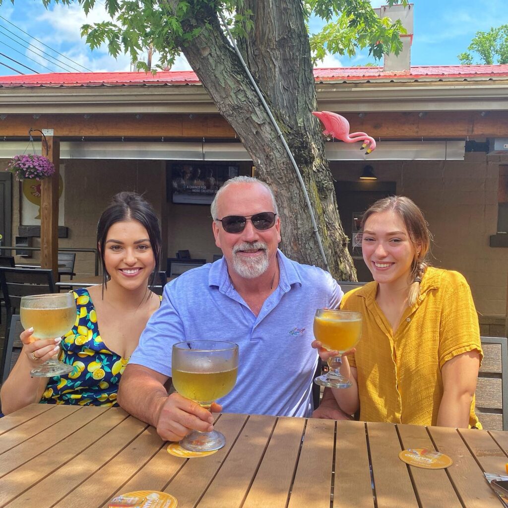 Two women sit on either side of a man. All three people have fishbowl glasses filled with a golden beverage on an outdoor patio.