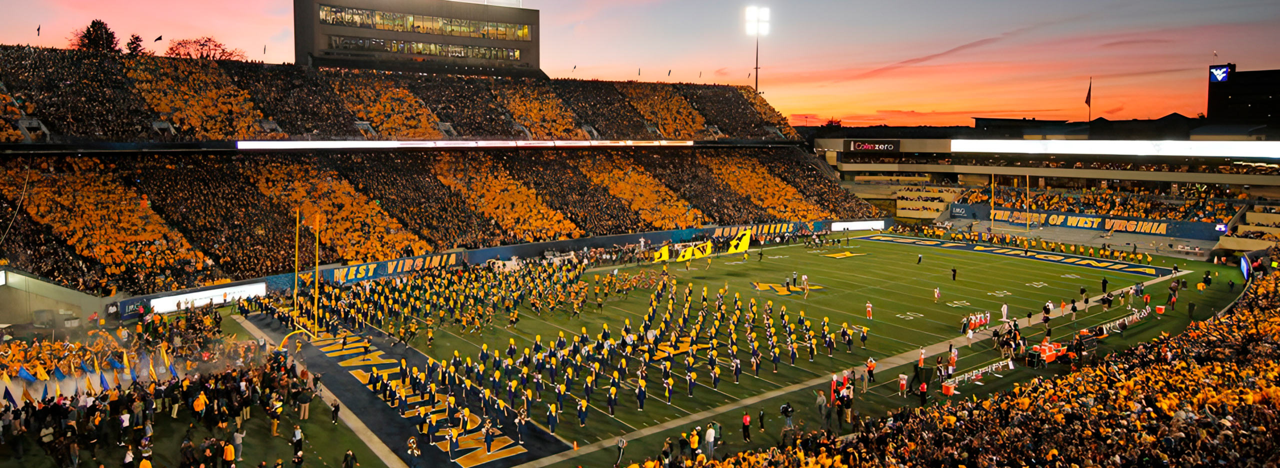 wvu-football-hero-2 A football stadium is striped into navy blue and gold sections. Football players are on the field. The stadium is full. There is a pink and orange sunset in the distance.