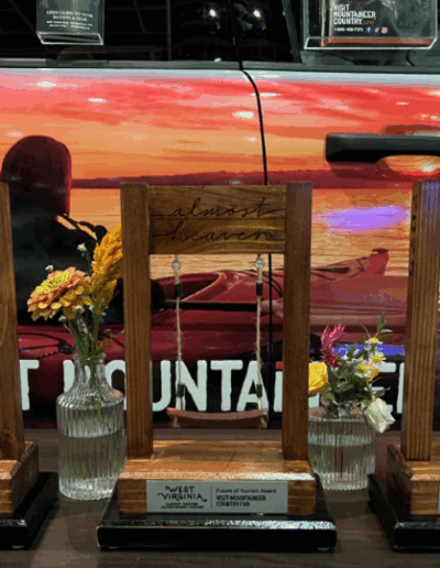 Three wooden awards in the shape of wooden swings are displayed i a lie in front of a bright, colorful vehicle with a logo that says "Visit Mountaineer Country" on the side. The awards say "Future of Tourism," Tourism Development, GeoWoodstock," and "Digital Innovation." In between the swings are two small bud vases with flowers.