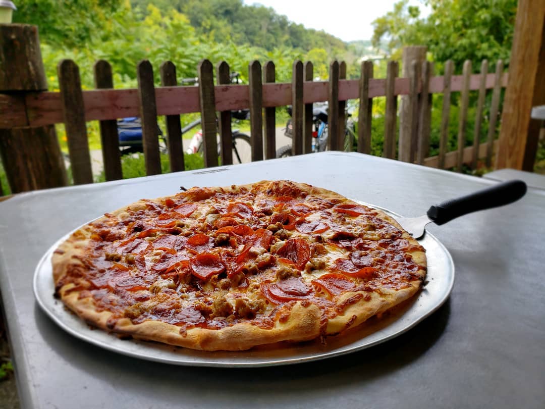 A pizza with pepperoni and other meats sits on a pan fresh out of the oven. It is on a tabletop, and there is a fence and greenery in the background.